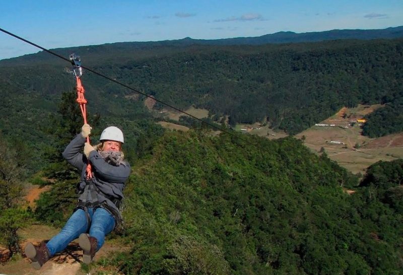 Foto da tirolesa K2mil na cidade de Rodeio, em Santa Catarina. Uma mulher olha para a foto enquanto desce a tirolesa. Ao fundo uma imensa vegetação. 