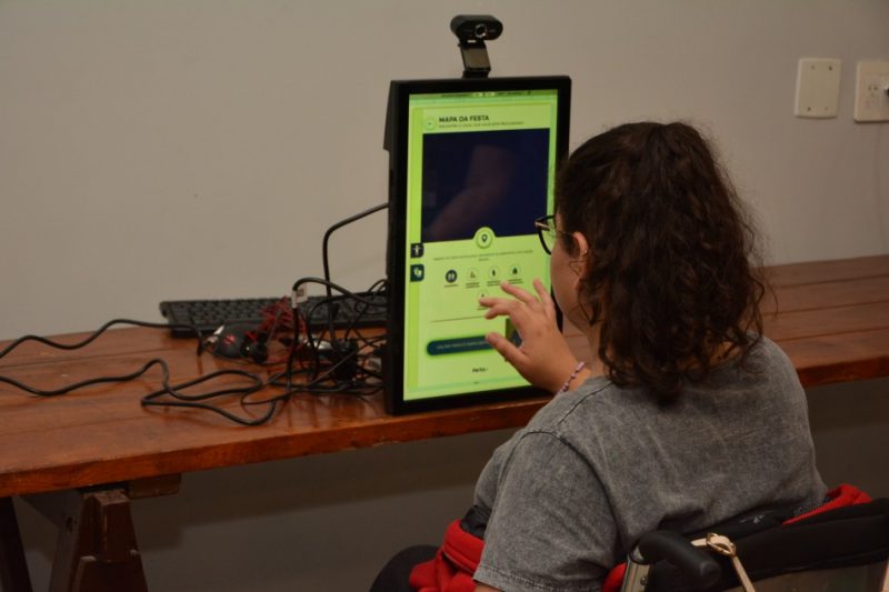 Moça em uma cadeira de rodas testando um toten tecnológico a ser usada na Oktoberfest Blumenau. Ela está de costas para a foto. Tem cabelos castanhos e ondulados e usa uma camiseta cinza. 