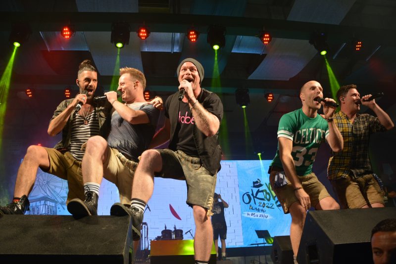 No palco, cinco homens com diferentes características físicas. Eles usam bermuda cor bege, camisetas e botas pretas. Eles estão cantando em uma apresentação na Oktoberfest Blumenau. 