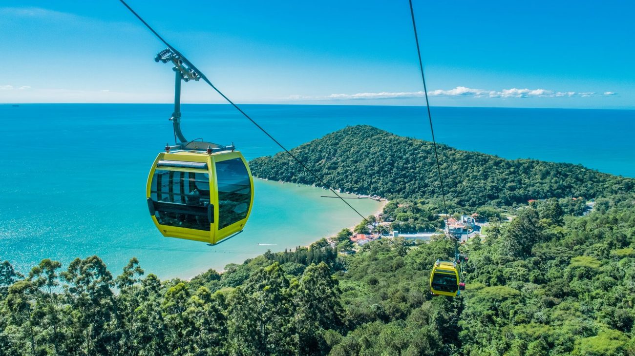 Em primeiro plano aparece uma cabine amarela do bondinho de Balneário Camboriú. Ele está passando por cima de um morro verde em direção à Praia de Laranjeiras, que se vê ao fundo, com um mar azul claro. 