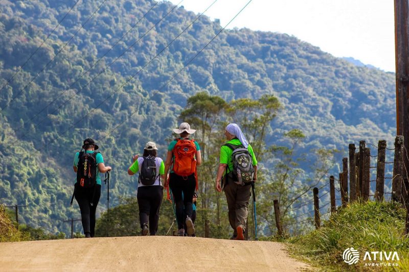 Foto mostra quatro pessoas de costas caminhando por uma estrada de terra na região do Vale Europeu. Elas estão com mochilas, bonés e cajados de caminhada. Ao fundo, aparece um morro verde.