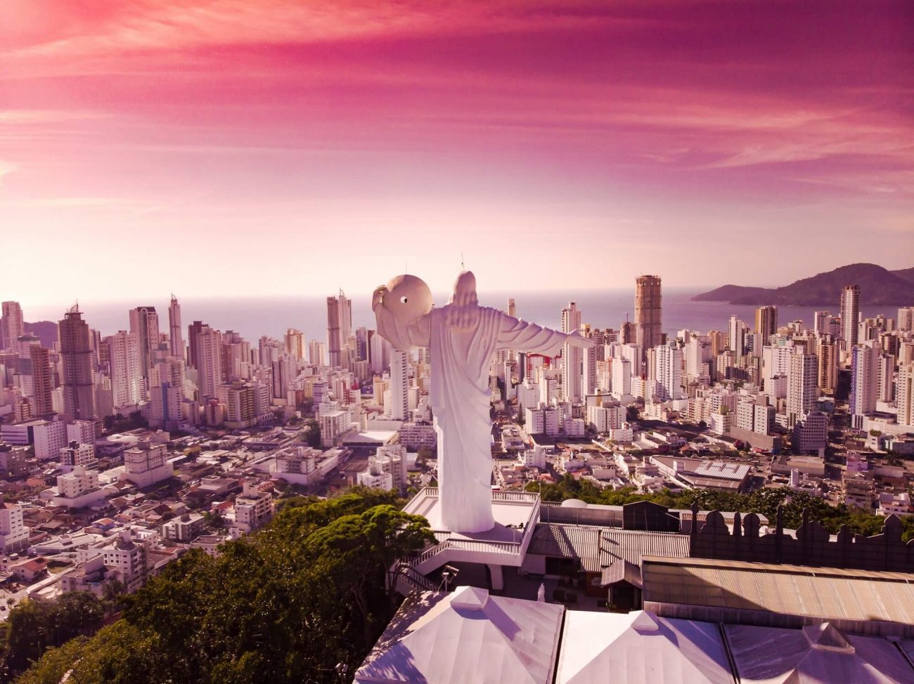 O Cristo Luz, em Balneário Camboriú, aparece de costas para a imagem. À frente dele, estão muitas casas e altos prédios e depois destes, o mar. 