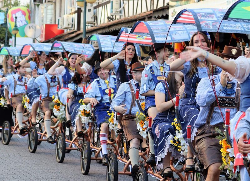 Foto de desfile na rua durante a Oktoberfest de Blumenau. Na imagem, uma bicicleta com várias assentos e coberturas, onde estão sentadas várias pessoas vestidos com um traje típico.