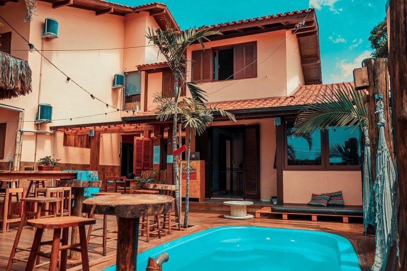 Imagem de um hostel na Barra da Lagoa em Florianópolis. A foto mostra aos fundos um sobrado na cor de salmão e janelas em marrom. Na frente, está um quintal. Aparecem algumas mesinhas e bancos de madeira e parte de uma piscina. Há palmeiras também. 