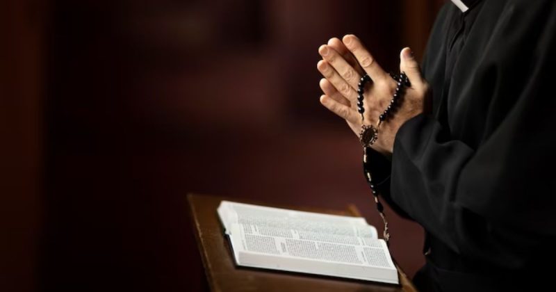 Mãos de padre com terço rezando e bíblia em púlpito 