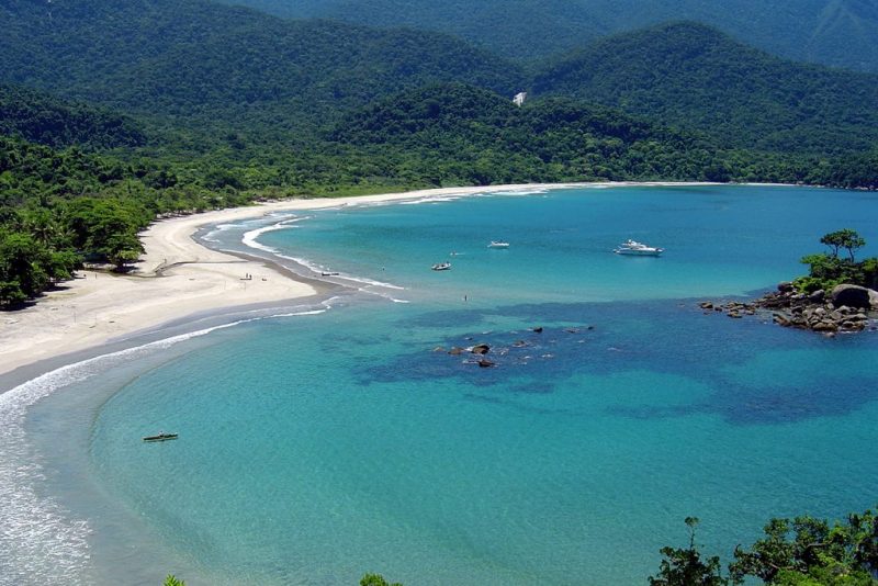 Foto de Ilhabela, com um mar azul claro, uma faixa de areia branca e morros verdes ao fundo. A cidade é destino turístico de cruzeiro saindo de Itajaí na temporada 2023-2024