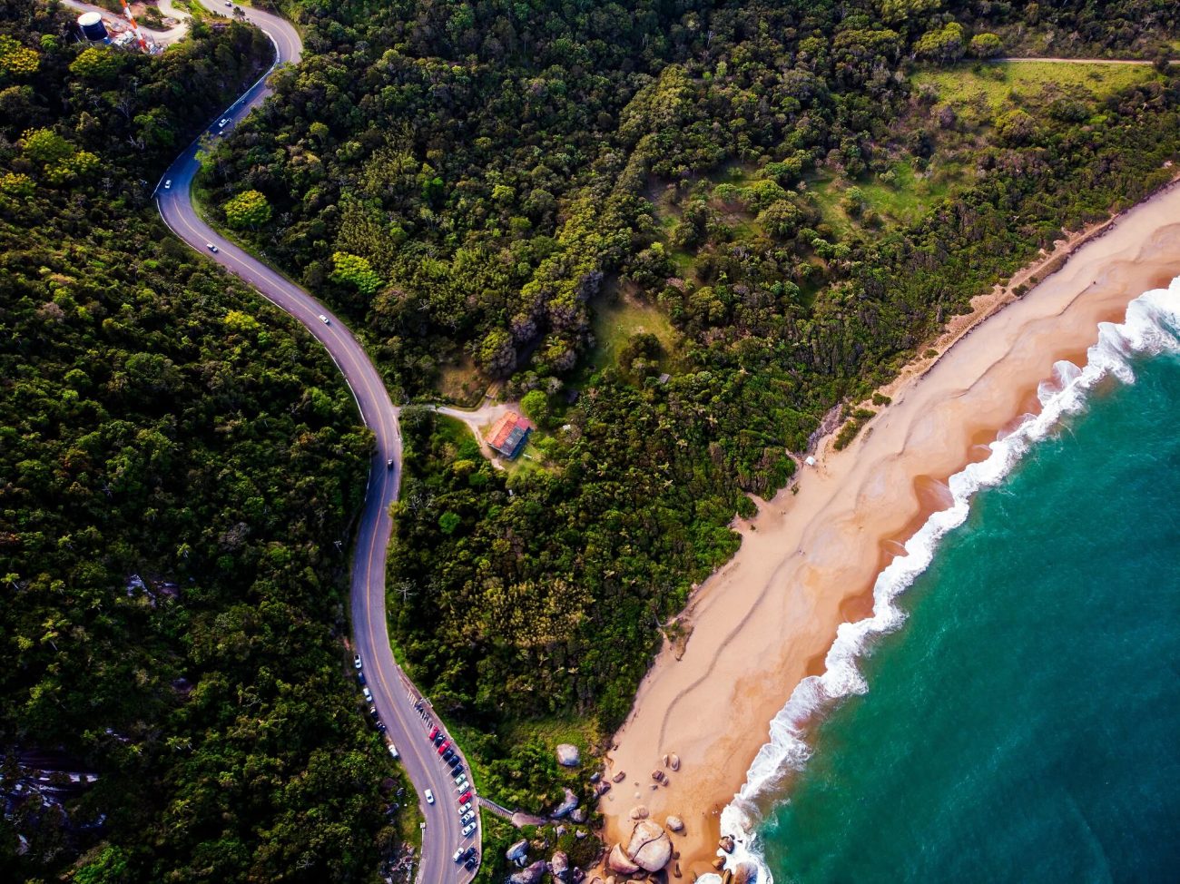 Foto aérea de um trecho da Rodovia Interpraias em Balneário Camboriú. A foto mostra uma imensidão verde e a rodovia de asfalto e algumas de suas curvas na parte esquerda da imagem. Na parte inferior direita aparece um trecho de areia e o mar de águas esverdeadas. 