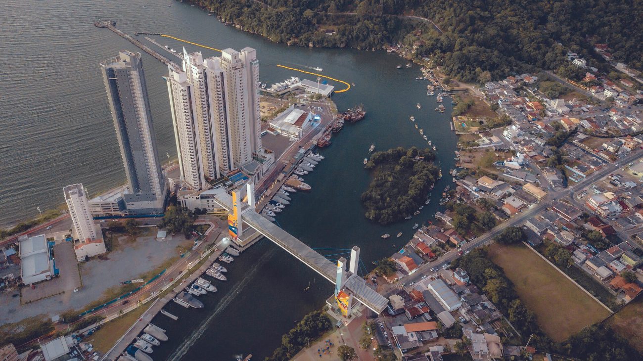 Foto aérea da Passarela da Barra em Balneário Camboriú. Ela é uma estrutura alta de concreto que passa por cima do Rio Camboriú, onde estão estacionados alguns barcos. De um lado do rio há grandes e modernos prédios. Do outro, casas baixas e uma paisagem mais verde. 