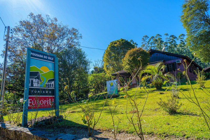 Foto mostra em primeiro plano uma placa turística do Vale Europeu. Ao fundo um jardim e uma pousada de madeira, na cidade de Benedito Novo.