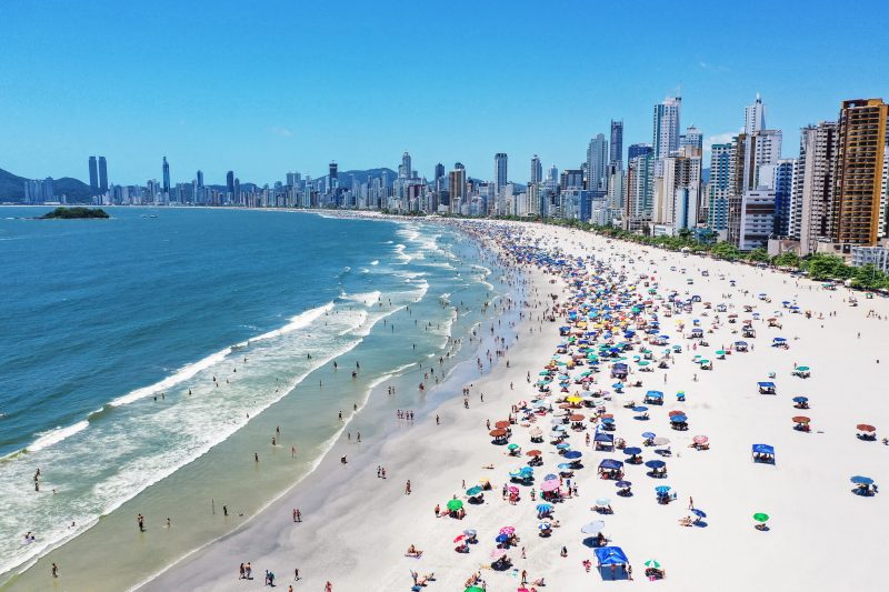 Foto da orla da Praia Central de Balneário Camboriú. À direita, a faixa de areia branca repleta de guarda-sóis coloridos. Ao fundo, os altos prédios da cidade. Do lado esquerdo, o mar azul. 