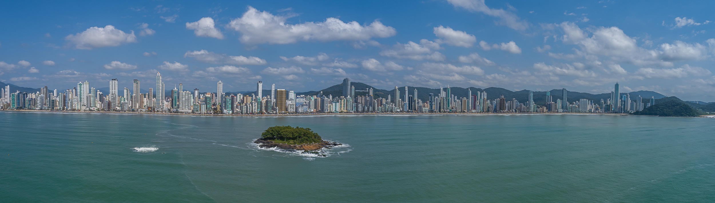 Foto panorâmica da Praia Central de Balneário Camboriú. Em primeiro plano aparece o mar e uma pequena ilha. Ao fundo estão os grandes prédios da cidade e atrás deles morros verdes. 