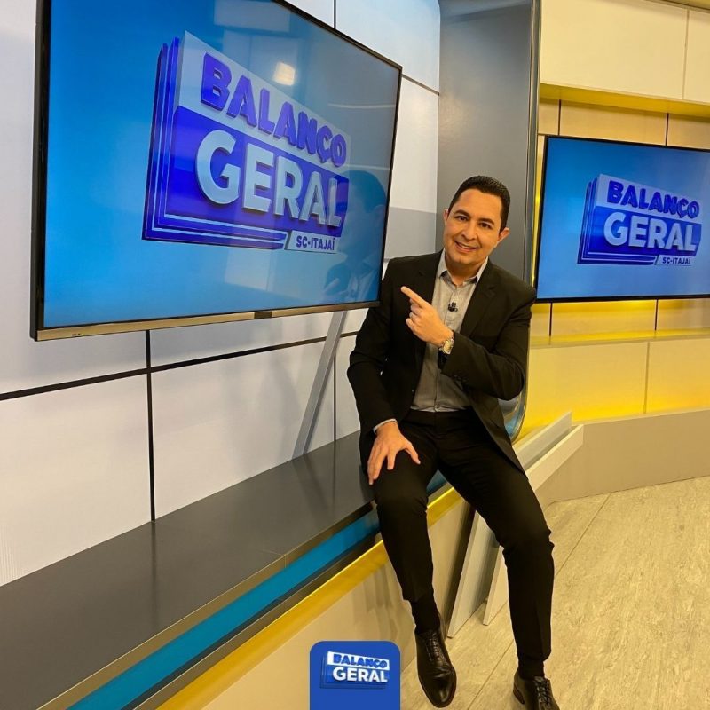 Calebe Moreno, apresentador do Balanço Geral Itajaí, aponta para uma das televisões do estúdio