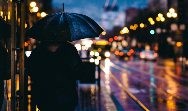 Pessoa com guarda-chuva em meio a rua molhada à noite, em centro iluminado