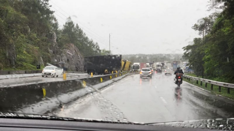 Foto mostra uma carreta tombada atrapalhando o fluxo do trânsito na pista. Registro foi feito de dentro de um carro, na foto é possível ver a pista molhada pela chuva.