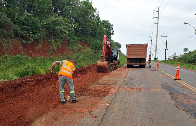 Homem trabalhando com uma enxada no canto do asfalto durante as obras do Contorno Viário Oeste, em Chapecó. Máquinas aparecem na pista removendo a terra do local 