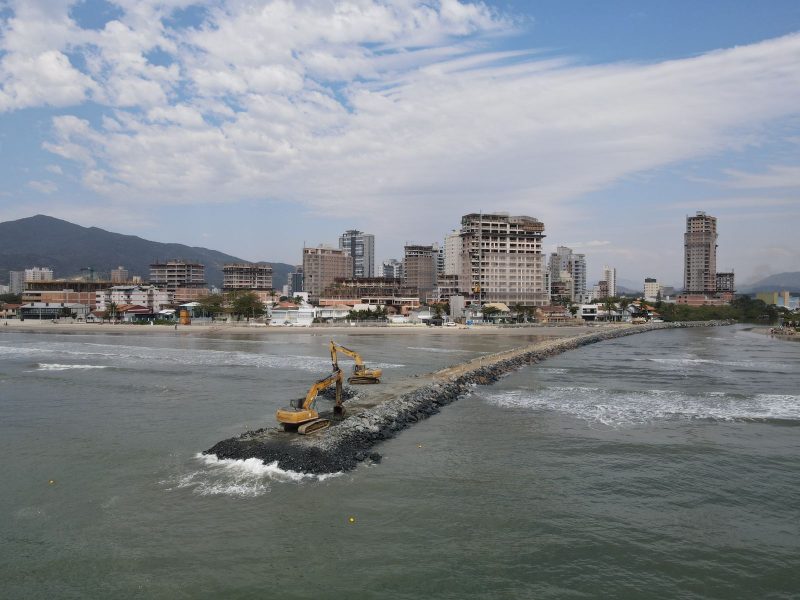 Imagem mostra construção do molhe sendo feita, com maquinário trabalhando na região em meio ao mar