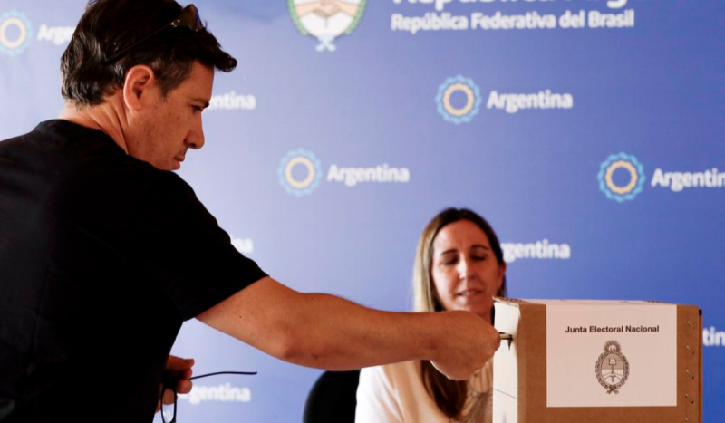 Eleitor inserindo cédula de voto em urna de papel. Ao fundo, um painel com a insígnia da Embaixada da Argentina no Brasil