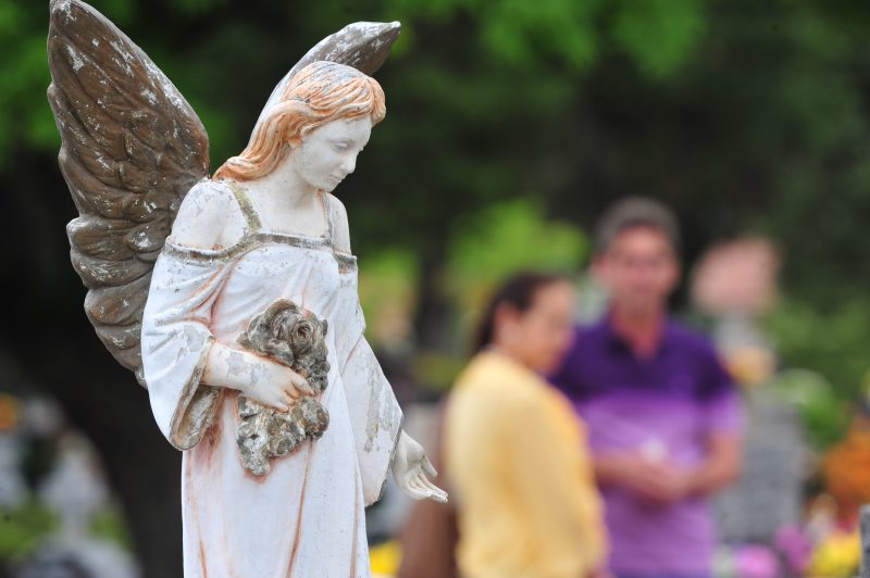 Estátua de anjo simbolizando o Dia de Finados em destaque; ao fundo, fiéis relembram entes que já partiram, em meio a cemitério