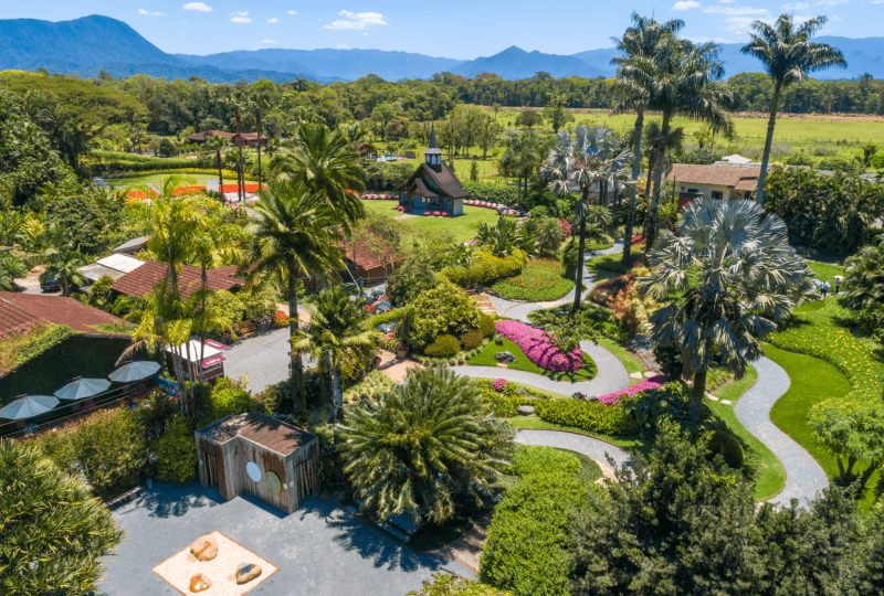 Foto aérea do Parque Hemero em Joinville. Em um ângulo de cima é possível ver árvores e um amplo jardim gramado, recortado em curvas, e com flores. Ao fundo a capela de madeira e no horizonte morros. 