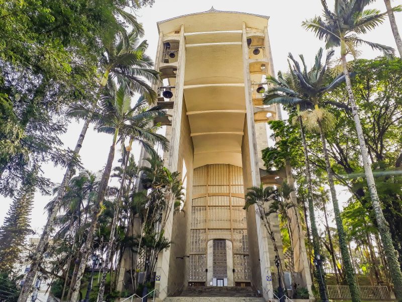 Foto do portal da Igreja Matriz de Brusque. A fotografia foi registrada em uma perspectiva de baixo para cima, dando mais imponência ao alto portal de concreto em cor bege. Na parte superior dele há dois sinos de cada lado. Ao seu redor, algumas palmeiras e outras árvores. 