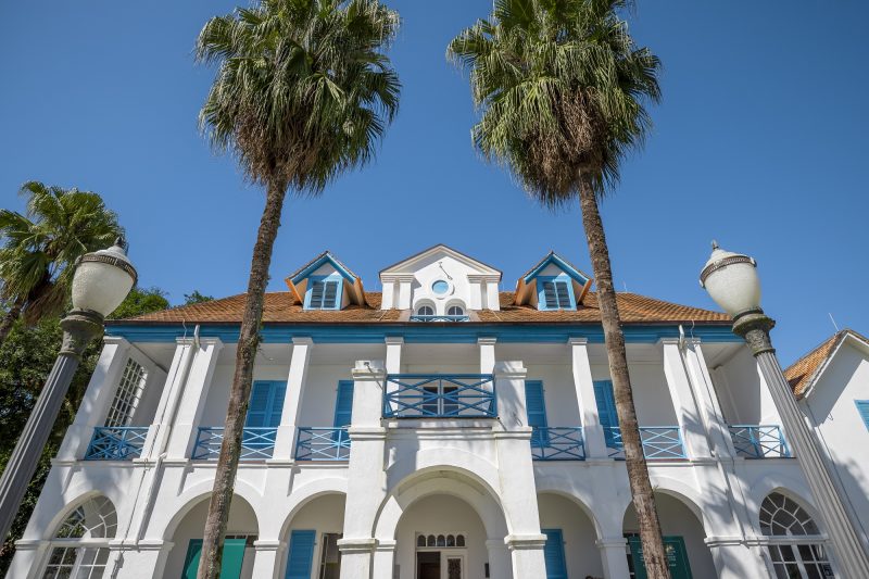 Foto do Museu Nacional da Imigração em Joinville. O museu é um casarão sobrado com paredes brancas e janelas, portas e parapeito de madeira azuis. A foto foi registrada em um ângulo de baixo para cima, que enquadrou nas laterais do casarão dois postes de luz e no centro do casarão duas palmeiras, que ficam no jardim do museu. 