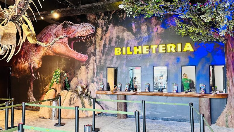 Foto da bilheteria do Parque Aventura Jurássica. Em primeiro plano aparecem organizadores de fila. Ao fundo, uma parede com pintura de pedras, árvore e um dinossauro com guichês de atendimento. No centro da parede, escrito em amarelo: Bilheteria. 