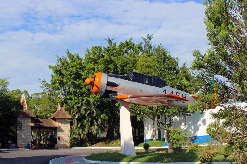 Foto da entrada do Parque Leopoldo Moritz em Brusque. No meio da foto e um pouco à direita aparece a réplica de um avião North American T-6D. Ele é cinza com vidros pretos e asas laranjas. Está erguido do chão por uma pequena torre de concreto. Ao redor dele há árvores e ao fundo aparece o portal de entrada do parque: duas torres de tijolinhos à vista com um vão no meio. 