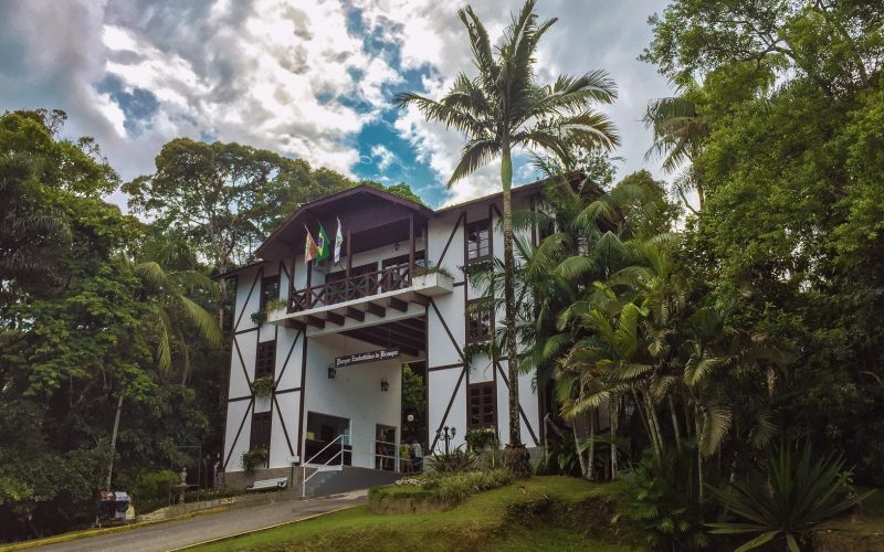 Foto do portal de entrada do Parque Zoobotânico em Brusque. Ele é um prédio de dois andares com um vão no meio. É branco com janelas e sacada marrom. Nas paredes da frente há ripas de madeira entrecruzadas, que imitam a técnica de construção germânica enxaimel. Ao redor dele há árvores. 