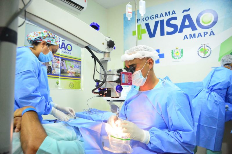 A surgeon operates on a patient's eyes.  He sits wearing a mask and with his face covered in surgical equipment.  A nurse helps with instruments, and behind them can be seen a panel with the logo of the Mias Visão program, which is analyzed after cases of blindness in patients infected with the fungus.
