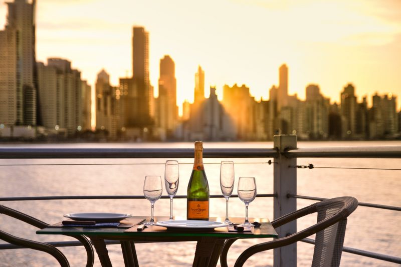 Foto da varanda do restaurante Porto Cabral em Balneário Camboriú. Em primeiro plano aparece uma pequena mesa redonda com taças, dois pratos e uma garrafa de vinho em cima. Ao redor, há duas cadeiras. Ao fundo aparece o mar e os arranha-céus de Balneário Camboriú, desfocados. 