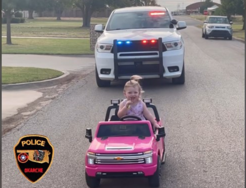 Menina de 2 anos dirige carrinho rosa enquanto escova os dentes. Atrás, uma grande viatura da polícia.