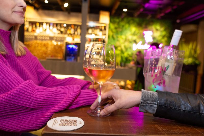 Foto mostra uma mesa de bar com duas taças de vinhos. Aparece metade do rosto de uma mulher, do nariz até a cintura. Ela é loira, tem cabelos na altura dos ombros e veste uma blusa de manga comprida rosa. À sua frente aparece o braço de um homem que segura a taça. Aparece apenas uma pedaço da manga da jaqueta de couro dele.