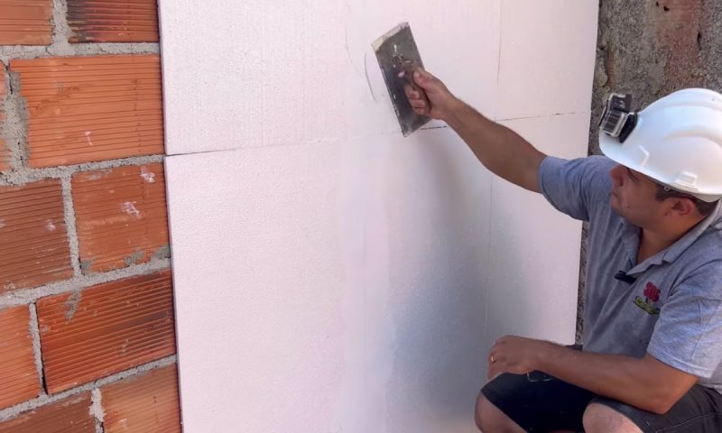 Pedreiro passa massa acrilica na placa de isopor colada na parede de tijolos