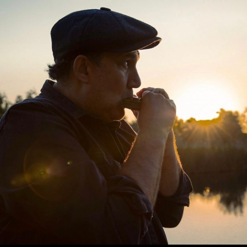 Imagem mostra o artista Alex Rossi tocando gaita com o por do sol ao fundo para promover o lançamento da sua nova Música