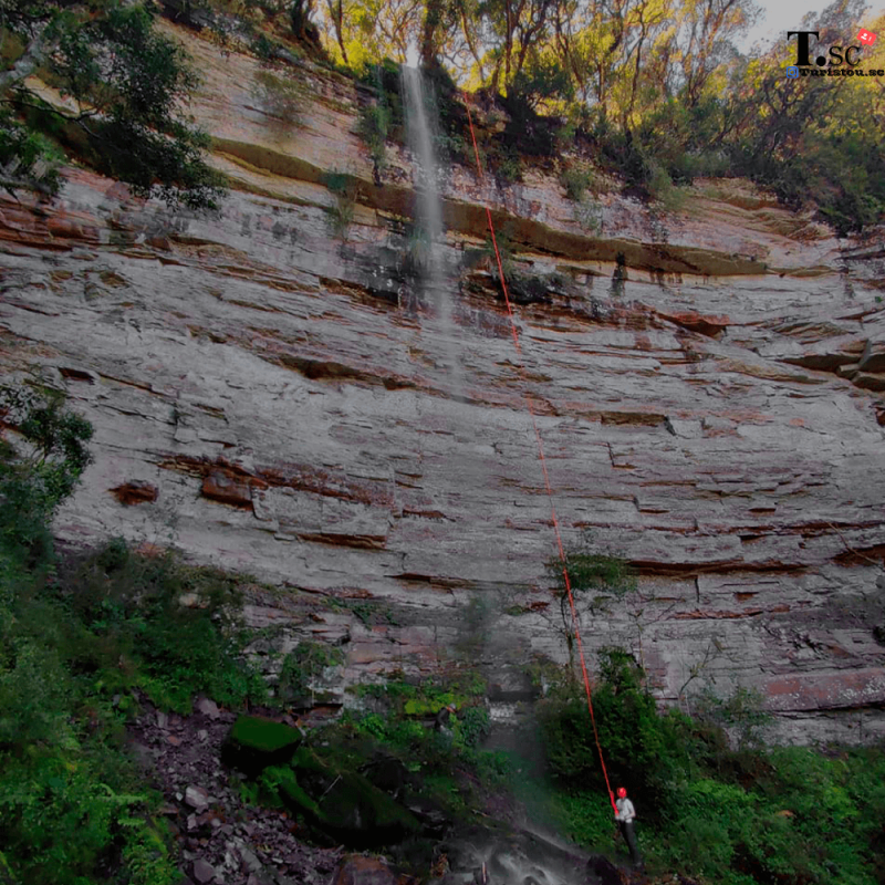 Foto de rapel na Cachoeira do Abraão, em Bom Retiro, na Serra Catarinense. Foto mostra um paredão rochoso com vegetação em cima e em baixo dele. Pelo paredão escorre uma fina cachoeira. Ao lado dela há uma corda laranja, presa a um homem que está no chão, ao fim da cachoeira. 
