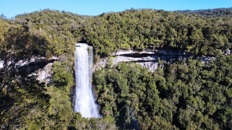 Foto da Cachoeira do Zinco, em Benedito Novo. A enorme e caudalosa cachoeira escorre em um paredão de pedras coberto pela vegetação e que cobre quase a totalidade da foto. Na parte superior, aparece uma faixa do céu azul. 