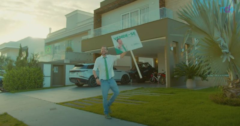 Imagem mostra corretor de imóveis com placa de vende-se em frente a casa de luxo em Balneário Camboriú, durante trailer de série "todo mundo odeia corretor de imóveis"