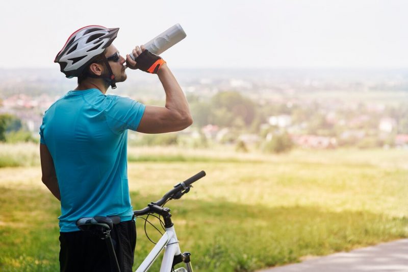 Ciclista segurando sua bicicleta e bebendo água em sua garrafa. Ele veste uma blusa azul, bermuda preta e capacete. Ao fundo e de forma desfocada aparece um campo gramado e algumas árvores 