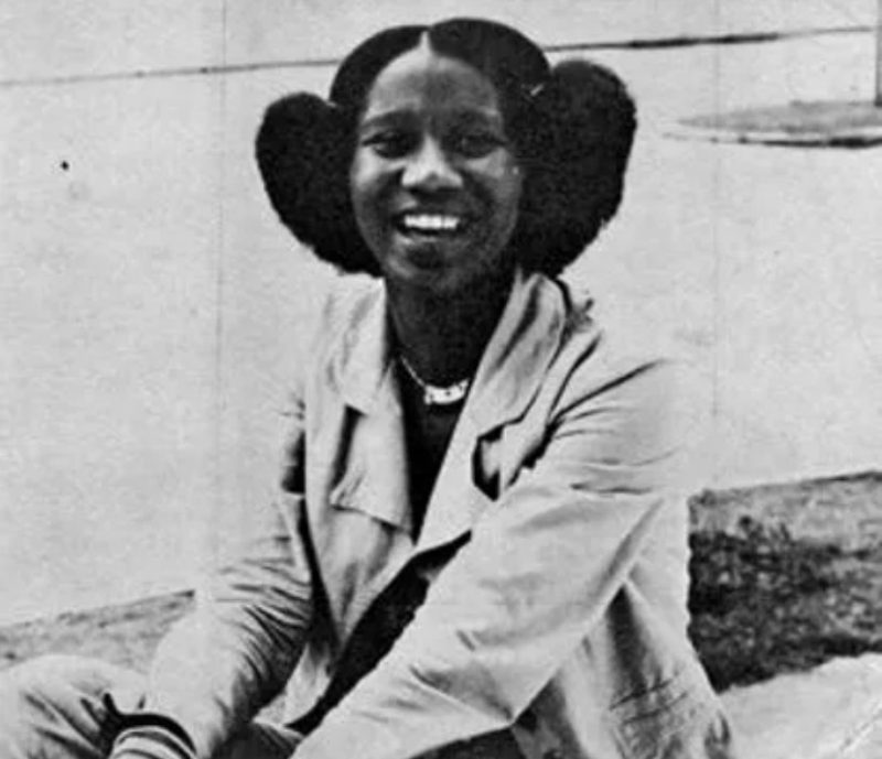 A foto traz a artista plástica Valda Costa, uma mulher negra, jovem e com sorriso intenso