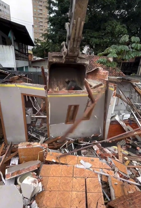 Foto mostrando uma escavadeira de dentro da cabine derrubando a estrutura de uma casa