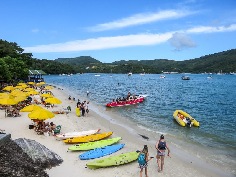 Foto da Ilha de Porto Belo. Do canto esquerdo ao centro aparece a areia da praia, com cadeiras e guarda-sóis amarelos e caiaques coloridos estacionados. Do centro ao canto direito aparece o mar com barcos e um banana boat. Ao fundo morros verdes. 