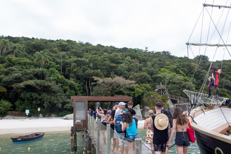 Foto de pessoas chegando à Ilha de Porto Belo. Do meio para o canto direito da foto está um trapiche de madeira por onde várias pessoas andam em direção à areia da praia, cercada de vegetação. Do lado direito dele está estacionado uma escuna com um homem fantasiado de pirata. 