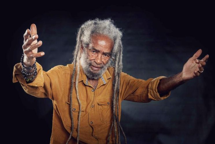 a foto traz o músico jamaicano Max Romeo, homem negro, de braços abertos. Ele é destaque da agenda cultural do final de semana em Florianópolis, quando fará show no domingo no John Bull Pub