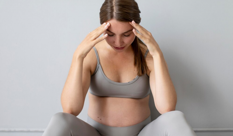 A pregnant woman sits with both hands on her forehead, as if she is worried.