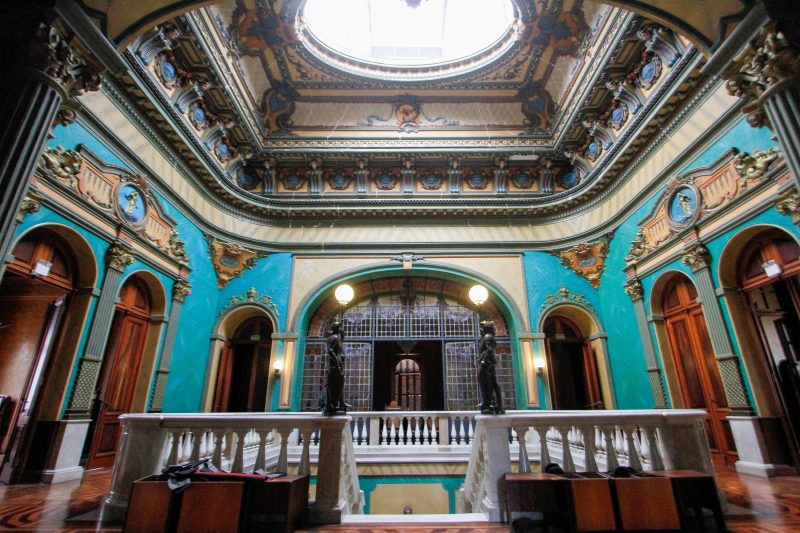 Foto interna do Palácio Cruz e Sousa, onde está instalado o Museu Histórico de Santa Catarina. No centro da foto aparece o primeiro degrau da escada que leva até o piso inferior. O vão, onde desce a escada é todo circundado por balaústres brancos. As paredes ao redor são pintadas nas cores azul e bege e são ricamente ornamentadas, até o teto. No teto, que é arredondado, há um círculo de vidro, por onde entra luz. Aparecem também seis portas de madeira. O chão também é de madeira e está envernizado. 
