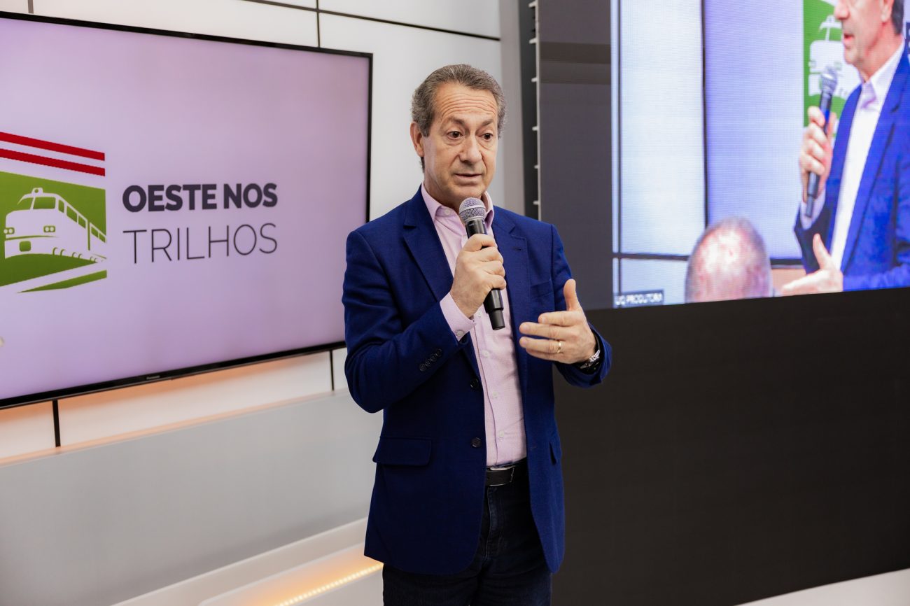 Senhor Caucasiano em pé, de blazer azul, com um microfone na mão e uma televisão com uma capa escrita Oeste nos Trilhos