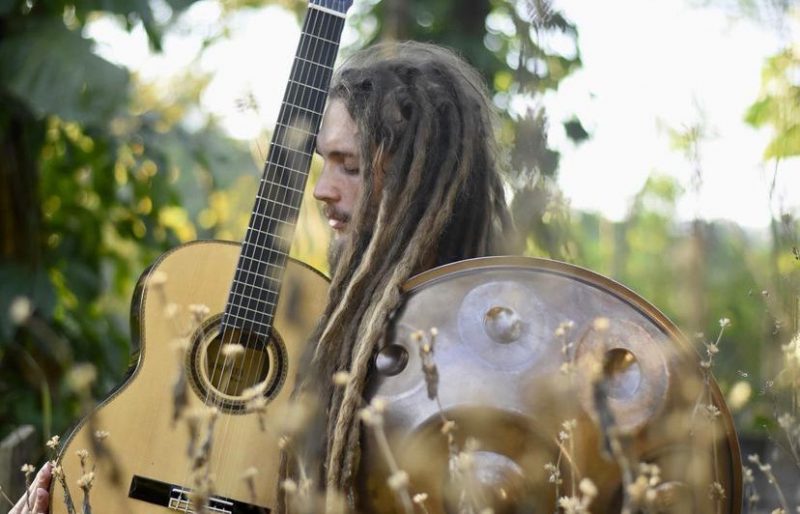 Um homem de cabelos compridos segura um violão. Ele se chama Pablo Vares e é destaque da agenda cultural