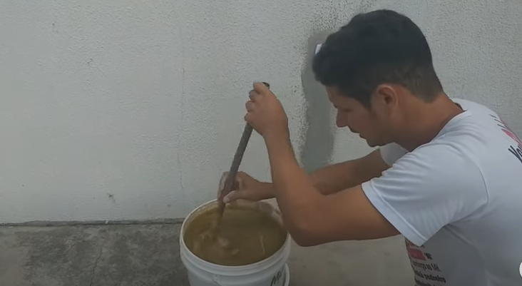 Pedreiro ensina a fazer a própria cor de tinta, para a pintura de casa; ele aparece mexendo com um tubo a tinta marrom dentro de um balde branco 