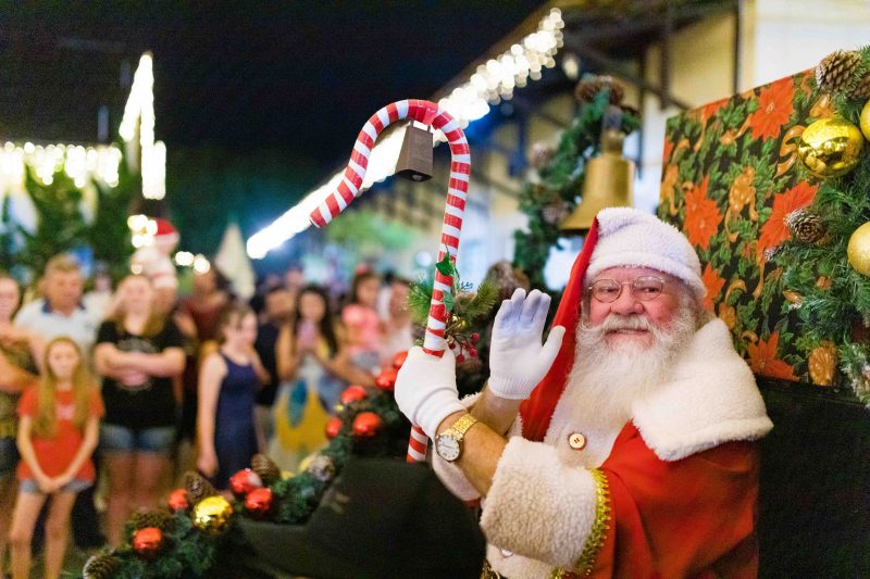 Foto de um homem idoso, de pele clara, óculos e barba grande e branca vestido de Papai Noel, sentado em uma poltrona. Ao fundo, pessoas desfocadas e parte do cenário do Centro Cultural de Pomerode também desfocado. É ali que ocorre o Natal, a última das festas em Pomerode. 