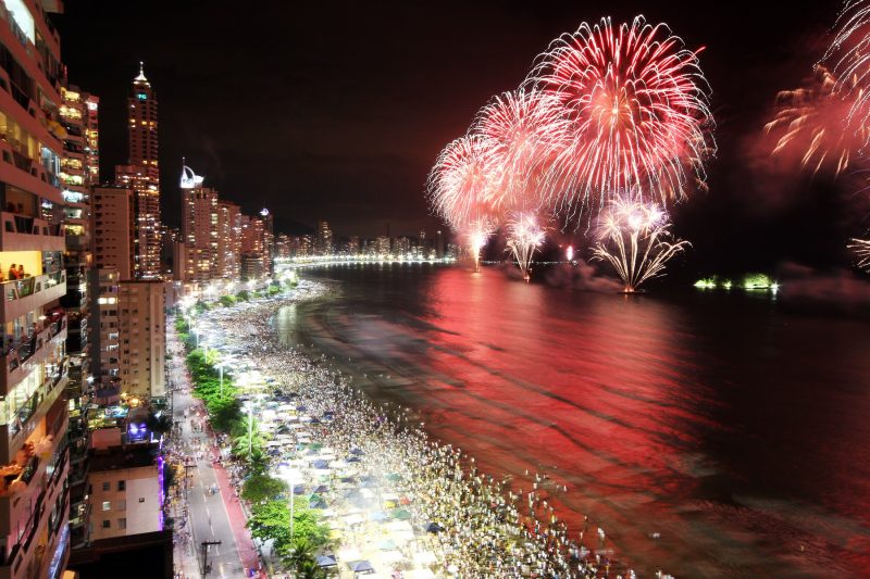 Show de fogos do réveillon na Praia Central de Balneário Camboriú; fogos de artifício explodindo ao fundo e iluminando o céu à noite, enquanto cidade está toda iluminada